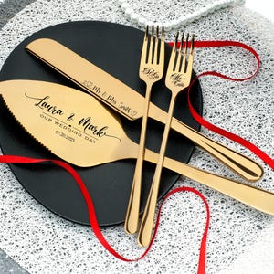 May include: Gold-toned wedding cake knife and two forks with engraved text. The knife has the text "Laura & Mark Our Wedding Day 07.20.2023" and the forks have the text "I Do" and "Me Too".
