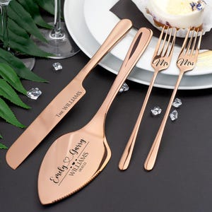 Puede incluir: Juego de cuchillo y servidor para pastel de boda en oro rosa con los nombres "Emily & Garry Williams" y la fecha "08.01.22" grabados en el servidor. Dos tenedores de oro rosa con "Mr" y "Mrs" grabados en los dientes.