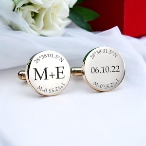 May include: A pair of gold-colored cufflinks with engraved details. One cufflink displays the initials "M+E" and the date "06.10.22" on the other. Both cufflinks have the coordinates "28°38'01.5"N" and "1°32'55.0"W" engraved around the edges.