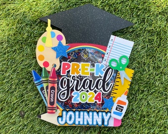 May include: A colorful pre-k graduation cap topper with a black graduation cap, a paint palette, crayons, scissors, a ruler, and a glue bottle. The topper says "Pre-K grad 2024 JOHNNY".