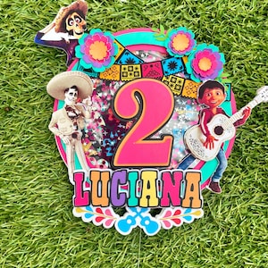 May include: A colourful cake topper featuring characters from the film Coco. The topper includes a pink number 2, a skeleton wearing a sombrero, a guitar-playing character, and the name "LUCIANA".