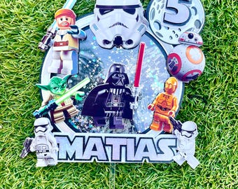 May include: A Star Wars themed cake topper with a black background and a silver glitter finish. The topper features Lego characters including Darth Vader, Yoda, a Stormtrooper, and C-3PO. The topper also includes a white Stormtrooper helmet, a Death Star, and the number 5. The topper says "MATIAS" in silver glitter letters.