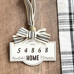 May include: White wooden Christmas ornament with a black and white striped ribbon bow. The ornament has the text "Christmas 2023" and the numbers "54868" with the words "Rice Lake HOME Wisconsin".