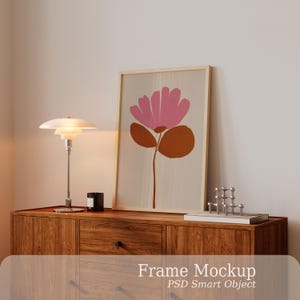 Puede incluir: Un marco de madera rectangular vertical que contiene una impresión de una flor rosa estilizada con hojas marrones sobre un fondo beige. El marco se apoya sobre un mueble de madera junto a una lámpara de mesa blanca de niveles y una pequeña escultura de metal plateado.