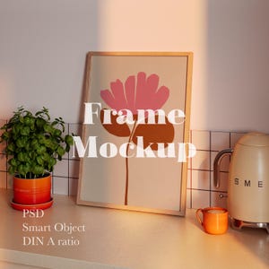 May include: A framed art print with a pink and brown floral design, labeled "Frame Mockup." A potted plant in an orange pot, an orange mug, and a cream-colored kettle are also visible. The image is set on a white countertop.