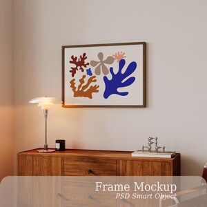 Può includere: Una cornice rettangolare orizzontale contenente una stampa astratta con forme organiche simili a coralli nei toni del rosso scuro, beige, rosa chiaro, blu e marrone. La cornice è montata su una parete liscia sopra un mobile in legno.