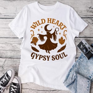 Puede incluir: Camiseta blanca con un diseño gráfico marrón con el texto "WILD HEART GYPSY SOUL". El diseño incluye la silueta de una persona, una luna creciente, estrellas, una rosa, plumas y cristales. Zapatillas negras y vaqueros en el fondo.
