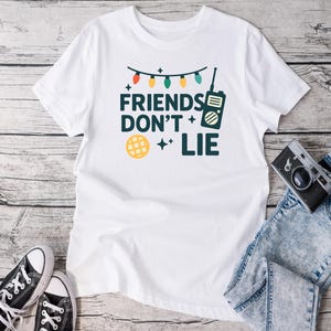 May include: White t-shirt with the text "FRIENDS DON'T LIE" in teal, with a string of colorful lights, a walkie-talkie, and a waffle graphic. Black and white sneakers and a camera are visible.
