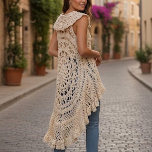 May include: A sleeveless, cream-colored crochet vest with a hood and a flowing, asymmetrical hem. The open-weave design reveals the wearer's blue jeans. The vest is a bohemian style garment.