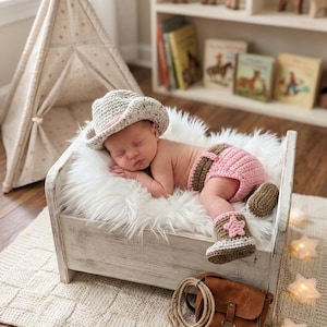 May include: A newborn baby wearing a crocheted cowboy hat, diaper cover, and boots, all in shades of beige, pink, and brown. The baby is lying on a small white bed with a faux fur blanket. A leather bag and rope are nearby.
