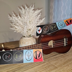 May include: A ukulele with five stickers on it. The stickers are black and white circles with different symbols. The ukulele is brown and has a light brown wooden neck. The ukulele is sitting on a wooden surface.