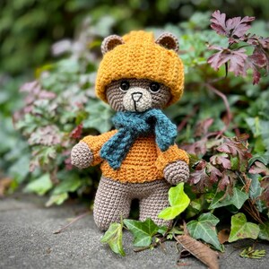 May include: A crocheted teddy bear wearing a mustard yellow hat, sweater, and a teal scarf. The bear has brown and tan coloring with black eyes and a light tan muzzle. The bear is standing in front of green and red foliage.