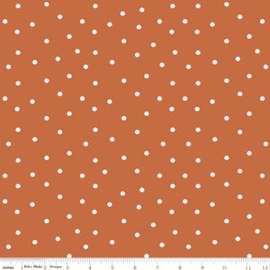 May include: A fabric with a repeating pattern of white polka dots on a rust-colored background. The fabric is measured in inches, with a ruler along the bottom edge. The text "Riley Blake Designs" is visible.