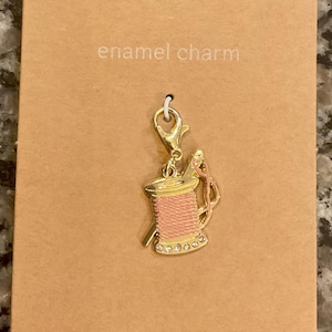 May include: Gold-colored enamel charm featuring a pink thread spool with a needle. The charm is attached to a gold clasp and is on a brown card. The card reads "enamel charm" and "JEWELRY MADE BY ME".