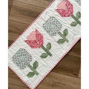 May include: A quilted table runner titled Posy Patrol featuring a repeating pattern of fabric flowers in red, pink, and light blue with green stems on a white background. The runner has a pink and white checkered border and measures 16.5" x 42.5".