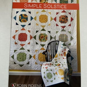 May include: A quilt pattern booklet titled "Simple Solstice" by Robin Pickens. The cover features a quilt with a sunflower pattern in shades of yellow, orange, red, green, and blue. The quilt is displayed on a chair in front of a white wall.