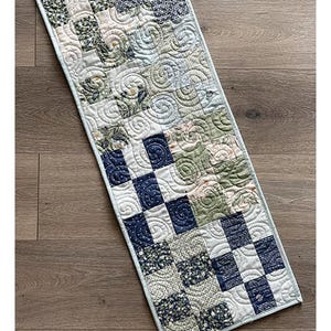 May include: A rectangular quilted table runner featuring a patchwork design of navy blue, sage green, and cream fabric squares with swirling stitch patterns. The text Benny Table Runner Project, Layer Cake & Fat Eighth Friendly, and finished size 12.5" x 54.5" are visible.