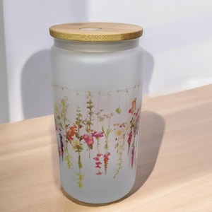 May include: A frosted glass jar with a wooden lid and a floral design. The jar has a white background with a design of colorful flowers hanging from strings.