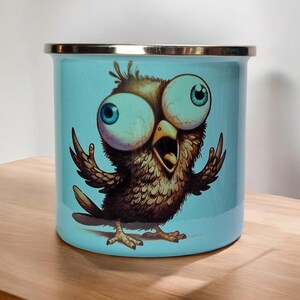 Può includere: Una tazza di smalto blu con un'illustrazione di un uccello marrone con grandi occhi blu e spalancati. L'uccello ha le ali spiegate e sembra sorpreso.