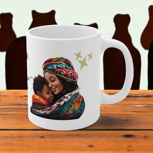 Può includere: Tazza in ceramica bianca con un'illustrazione colorata di una madre e un bambino che si abbracciano. La madre indossa un turbante colorato e il bambino una sciarpa colorata. Sullo sfondo ci sono stelle dorate.