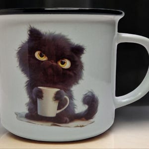 Op de afbeelding: Een witte keramische mok met een zwart handvat. De mok heeft een cartoon zwarte kat die een witte mok vasthoudt. De kat heeft gele ogen en een chagrijnige uitdrukking.