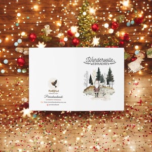 May include: A white card with a watercolor illustration of a winter scene with a Santa Claus figure and a deer. The text "Wundervolle Weihnachten" is printed in black at the top of the card. The card also features a logo for "Parissahandmade" with a bird graphic and the website address.