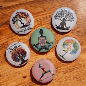 May include: Six round, colorful, printed buttons with inspirational messages and images of trees and yoga poses. The buttons feature phrases like "Embrace Stillness" and "Be an Optimist".