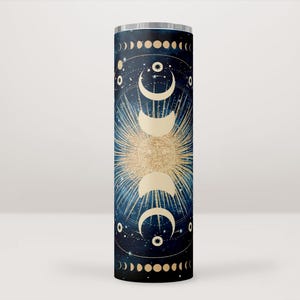 Pode incluir: Um copo de aço inoxidável com um design celestial. O copo apresenta um fundo azul escuro com detalhes dourados e um design de fases lunares brancas. As fases lunares estão dispostas em um círculo ao redor de um estouro de sol central.