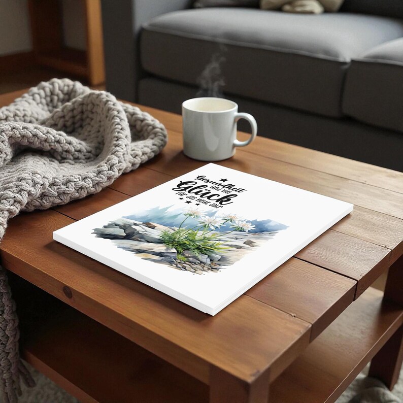 May include: A white canvas print featuring a watercolor illustration of a mountain scene with white flowers and the text "Gesundheit und Gl&uuml;ck f&uuml;r das neue Jahr". A gray mug with steam rising sits next to the print on a wooden table.