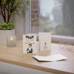 May include: A white card with a red and green illustration of Santa Claus holding a snowman. The text on the card reads "Frohe Weihnachten".