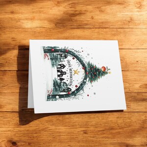 K&ouml;nnte beinhalten: Eine wei&szlig;e Weihnachtskarte mit einem gr&uuml;nen Weihnachtsbaum und einer schwarz-wei&szlig;en Illustration von H&auml;usern unter einem Torbogen. Der Text "Frohe Weihnachten" steht auf der Karte.