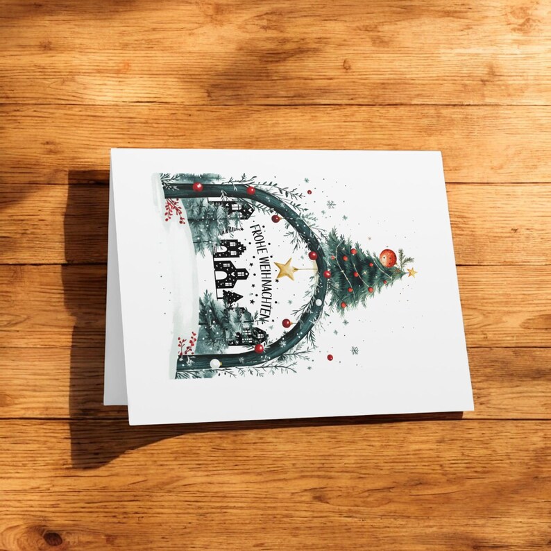 K&ouml;nnte beinhalten: Eine wei&szlig;e Weihnachtskarte mit einem gr&uuml;nen Weihnachtsbaum und einer schwarz-wei&szlig;en Illustration von H&auml;usern unter einem Torbogen. Der Text "Frohe Weihnachten" steht auf der Karte.