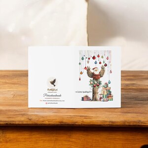 May include: A Christmas card featuring a cartoon bird wearing a Santa hat and holding a Christmas tree ornament. The bird is perched on a tree trunk with presents stacked underneath. The card has a white background with colorful Christmas ornaments hanging from the top. The text "Sch&ouml;ne Weihnachten" is written on the card.