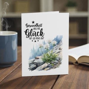 May include: A white greeting card with the German text "Gesundheit und viel Gl&uuml;ck f&uuml;r das neue Jahr" in black script. The card features a watercolor illustration of white flowers growing on a rocky mountainside. A blue mug and open book are in the background.