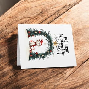 May include: A white cat wearing a red Santa hat sits under a Christmas wreath with ornaments and a gold star. The text "Frohliche Weihnachten" is written in black on the white card.