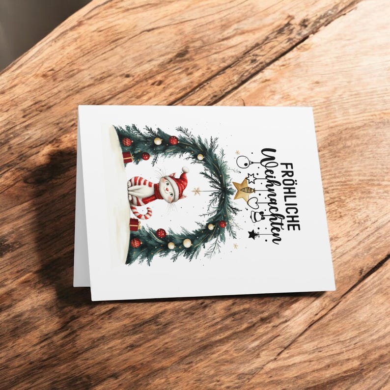 May include: A white cat wearing a red Santa hat sits under a Christmas wreath with ornaments and a gold star. The text "Frohliche Weihnachten" is written in black on the white card.