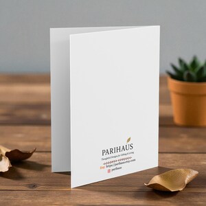 May include: A white folded greeting card with the text "PARIHAUS Thoughtful Designs for Gifting & Living" and a website address. The card is on a wooden surface with a small potted plant and a dried leaf.
