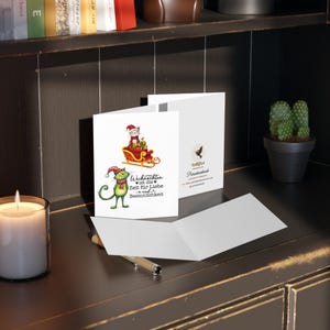 May include: A Christmas card featuring a green cat with a red Santa hat and a white cat riding in a red sleigh. The card has the text "Weihnachten ist die Zeit f&uuml;r Liebe und Besinnlichkeit" which translates to "Christmas is the time for love and contemplation".