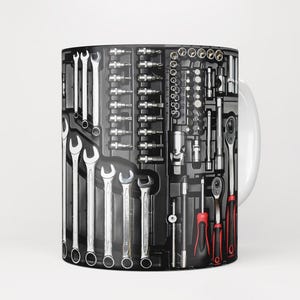 Könnte beinhalten: Eine weiße Keramiktasse mit einem schwarzen und silbernen Design aus verschiedenen Werkzeugen, darunter Schraubenschlüssel, Schraubendreher und Steckschlüssel.