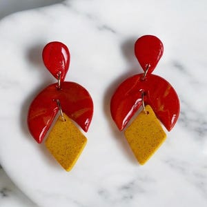 May include: A pair of dangle earrings with a red and yellow geometric design. The earrings feature a teardrop-shaped red piece, a half-circle red piece with yellow swirls, and a diamond-shaped yellow piece with glitter.