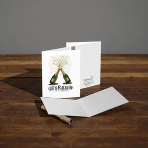 May include: White greeting card with a design of two champagne bottles clinking, with gold glitter and the words "Guten Rutsch". The card is open, revealing a blank interior, and is on a wooden surface.