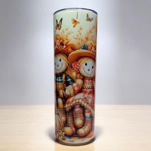 Pode incluir: Um copo branco com um design temático de outono que apresenta dois espantalhos sorridentes com chapéus de palha e botas marrons. O fundo é amarelo claro com detalhes marrons e verdes. Há borboletas nos cantos superiores do design.