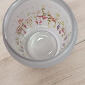 May include: A clear glass cup with a frosted exterior and a floral print on the inside. The flowers are in shades of pink, purple, and yellow.