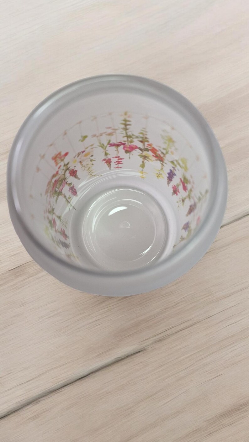 May include: A clear glass cup with a frosted exterior and a floral print on the inside. The flowers are in shades of pink, purple, and yellow.