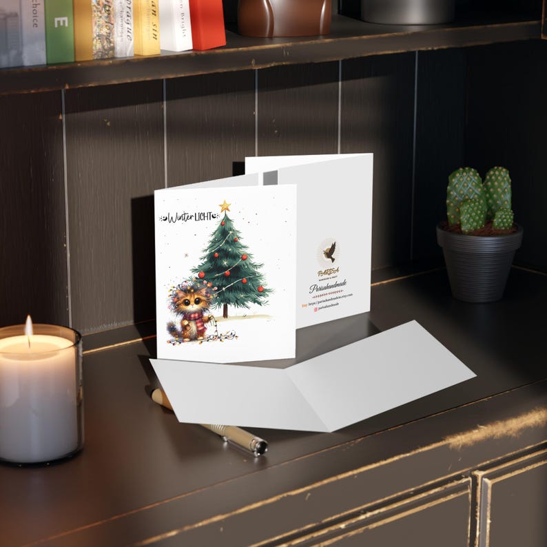 May include: A white card with a cute cartoon cat decorating a Christmas tree. The card has the text "Winter Lichter" on it. The card is folded in half and is sitting on a wooden surface.