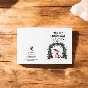 May include: A white Christmas card with a festive wreath and a cat wearing a Santa hat. The text "Fr&ouml;hliche Weihnachten" is printed on the card.