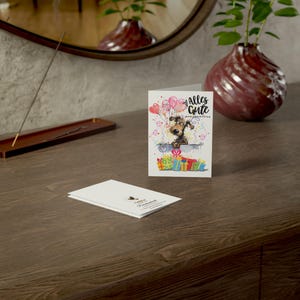 May include: A white card with a cartoon dog peeking over a pile of colorful presents. The card says "Alles Gute zum Geburtstag" in black text.