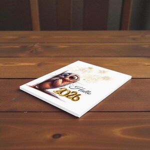 May include: A white rectangular card featuring a cartoon owl with large eyes and brown and gold coloring. The card has the text "Hallo 2026" in gold and black. Dandelion-like graphics are in the background.