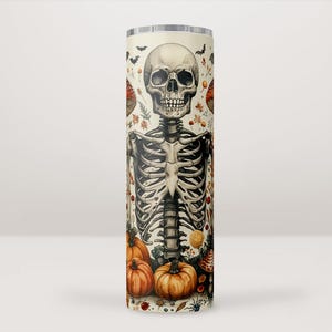 Puede incluir: Un vaso alto de color crema con una ilustración detallada de un esqueleto humano. El diseño incluye calabazas, murciélagos y follaje de otoño. El vaso es un producto con temática de Halloween.