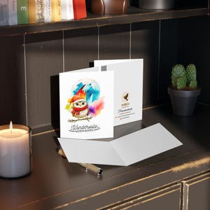 May include: A folded greeting card with a watercolor illustration of an owl wearing a red hat and scarf. The owl is perched on a branch with a colorful background. The text on the card reads "Wunderwulle. Weihnachten."